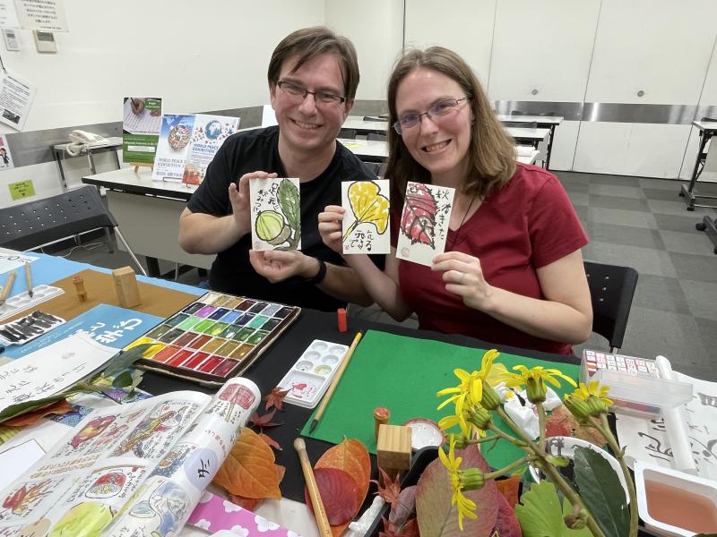 Amy and Andy are showing off their first Etegami artworks. They are holding up three painted postcards. Andy is holding up a painting of an acorn on a branch with a leaf. Amy is holding up two postcards. The first one is a painting of a golden ginkgo leaf. The second one is a painting of a red fall leaf. On the table in front of them is a gansai paint palette with 35 different colors, paint brushes, and various plants and flowers that they used as inspiration for their paintings. They appear in a classroom setting with various tables and chairs in the background.
